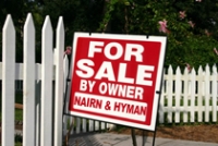 For Sale sign on lawn of a small home, in front of a white picket fence. 